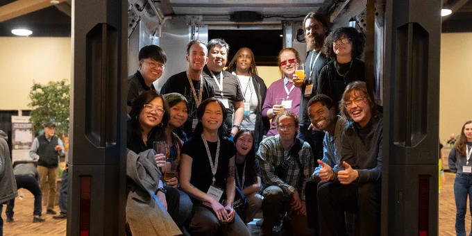 A group of smiling people posing inside a van at a conference, holding drinks and looking at the camera.