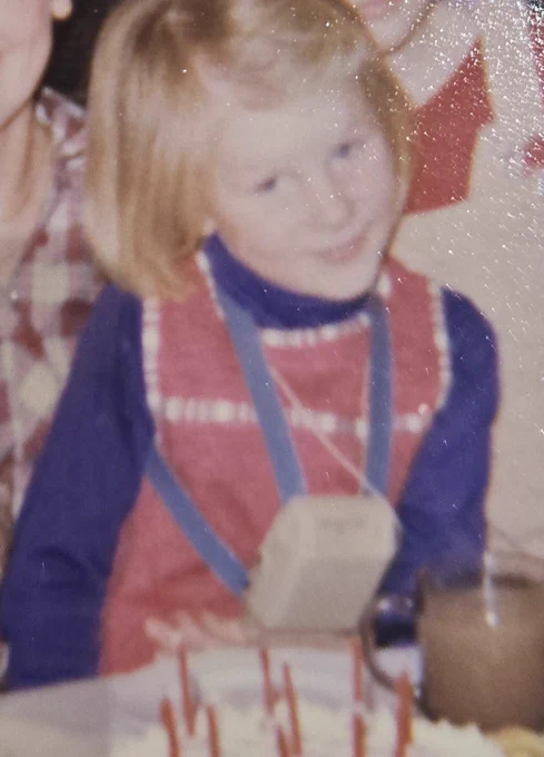 ID: a grainy photo of Jo as a child seated at a birthday party, she is wearing a boxy phonic ear (hearing aid). She has shoulder length blonde hair and is wearing a red dress and blue top
