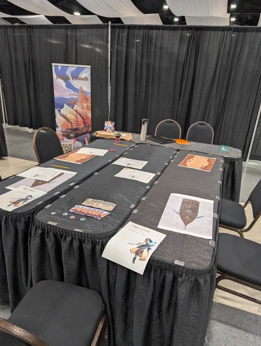 A table for High Winds RPG by Scale Bound Games at TAGCon in TriCities, WA. Three black tables are laid out with colorful pirate art, combat maps, and business cards. 