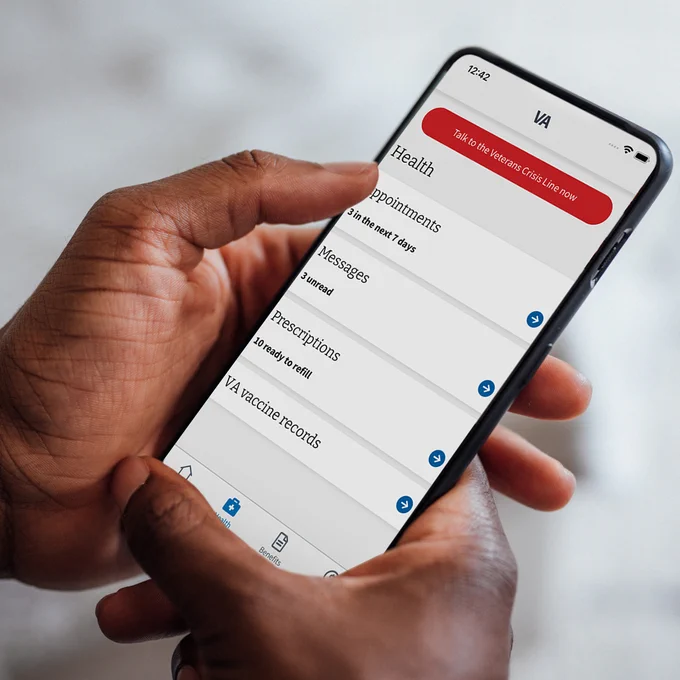 A person holding a smartphone displaying the VA: Health and Benefits app, which shows options for appointments, messages, prescriptions, and vaccine records. A red banner at the top provides a link to the Veterans Crisis Line.