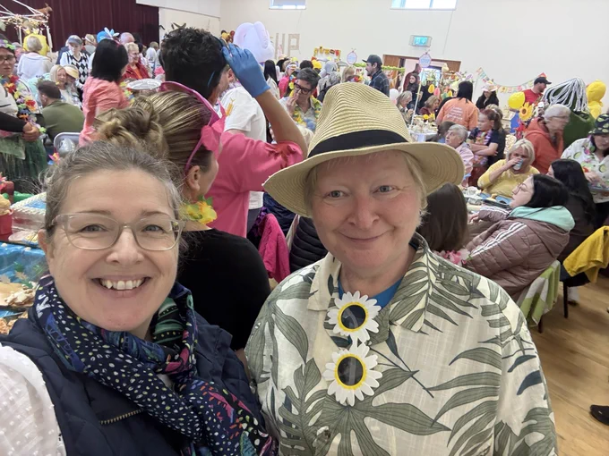 Kellie Armstrong MLA with Cransformation Lisa at the tea party to raise funds for the Air Ambulance. In the background are about 100 people in brightly coloured clothes, seated and standing