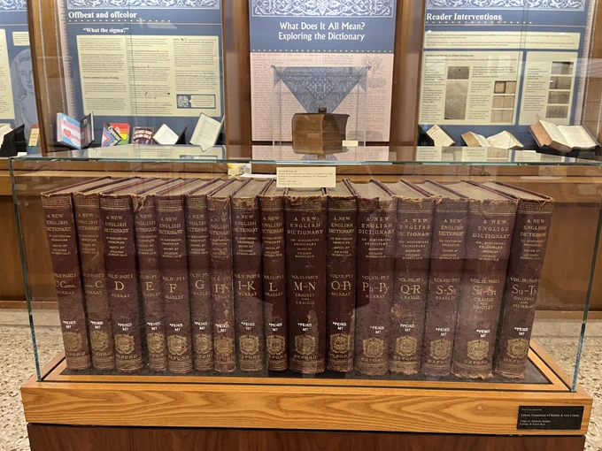 In the foreground 16 volumes of the New English Dictionary. In the background glass cases with posters on topics Offbeat and Offcolor; What does it all Mean, Exploring the Dictionary; Reader Interventions.