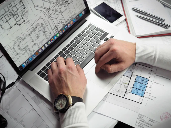 A person's hands are visible typing on a MacBook keyboard, which displays architectural blueprints. Nearby, there are architectural plans laid out on a table, a smartphone, and pens. The setting suggests an office environment focused on design or planning.