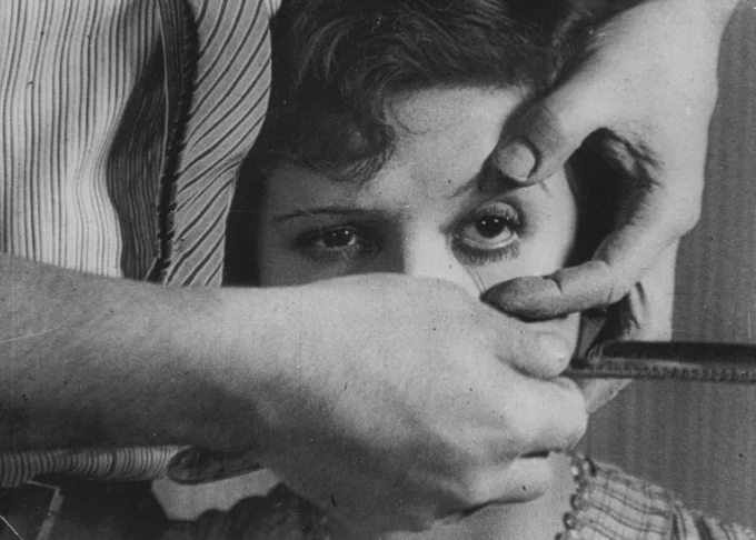 A black and white still of a woman sat down. A person stands behind her, holding her eye open, and holds a razor in their hand.