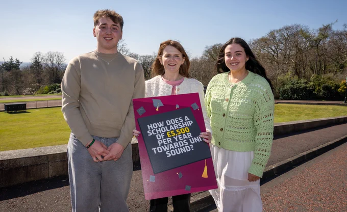 Economy Minister Dr Caoimhe Archibald with Taylor McGrath-McAllister, a second-year Natural Sciences student at Homerton College, University of Cambridge and second year student Elizabeth Poucher, who is studying Medicine at Queen’s University Belfast, both of whom are benefitting from a Scholarship. Minister is holding a graphic with the wording How does a Scholarship of £5,500 per year towards Uni sound?