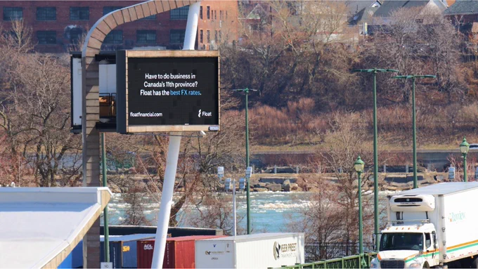 Billboard by Float displaying a message about doing business in Canada, with a river and industrial buildings in the background.