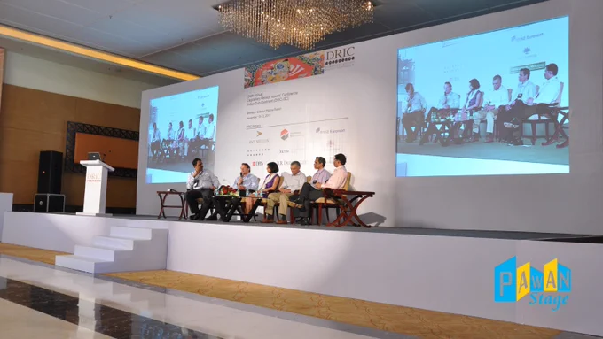 People gathered on a stage at Radisson Blu Udaipur, in front of a large screen displaying visuals for a conference event.