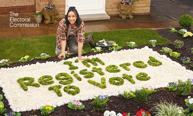 A young disabled woman is in her front garden, crouching over a flower bed. In the flower bed she has spelt out “I’m Registered To Vote” in grass.