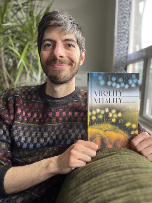 A smiling man (me) wearing a lovely sweater, holding a book he is evidently very proud of. The book is titled Virality Vitality, and has a blue and orange cover, with an image of a cell being invaded by ghostly bacteriophages