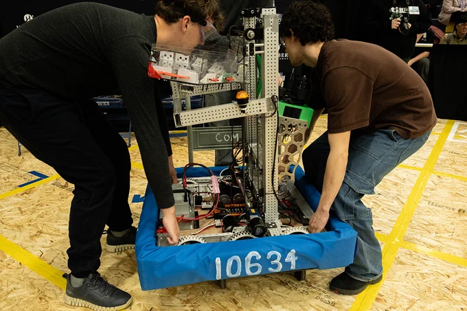 Two people carry a robot by its base, which has the number 10634 printed on it.

