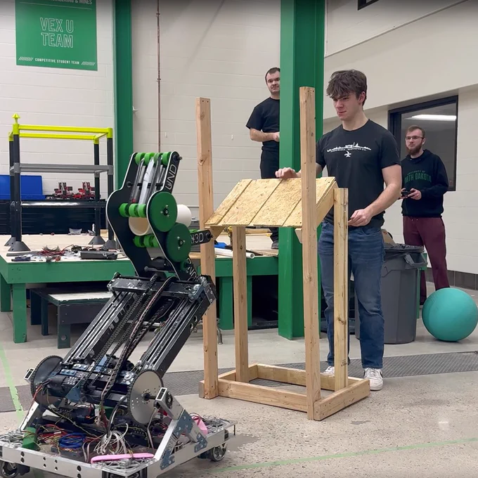A group of students demonstrates the functionality of a UND student-built robot that has a head that looks somewhat like a dinosaur. The robot is on wheels and sports UND-green accents.