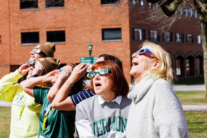 Group of people wearing eclipse glasses looking upwards with expressions of awe, standing in front of a brick building on a sunny day.
