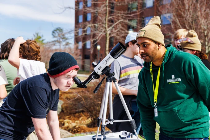 A person looks through a telescope during an outdoor event with guidance from a staff member wearing a green sweatshirt. Other attendees are visible in the background.