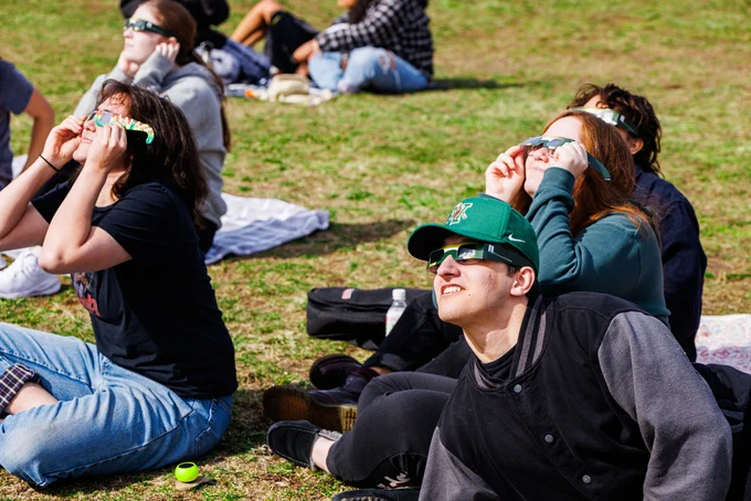 Group of people sitting on grass looking up at the sky wear solar eclipse sunglasses.