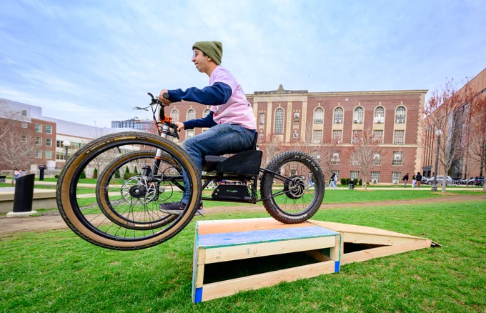 Grainger College of Engineering student riding an adaptive three-wheeled E-bike over a wooden ramp on the University of Illinois' engineering quad with a large brick building in the background.