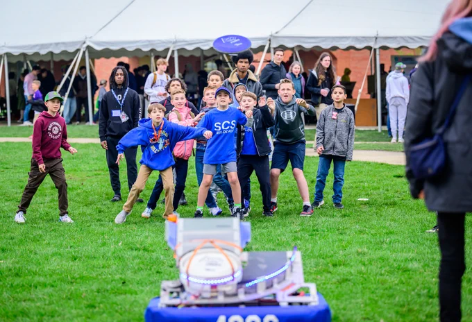 A group of enthusiastic middle-school-age students children wait in anticipation as a blue Frisbee-type disc is being hurled at them by a robotic device on the grass of the University of Illinois' engineering quad  Adults and tents are visible in the background.

