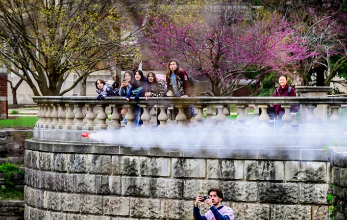 Group of people standing outdoors against a stone wall watching a small, orange model rocket whiz bu with a stream of white smoke trailing behind. In the foreground, a Grainger engineering student captures the launch on a camera.
