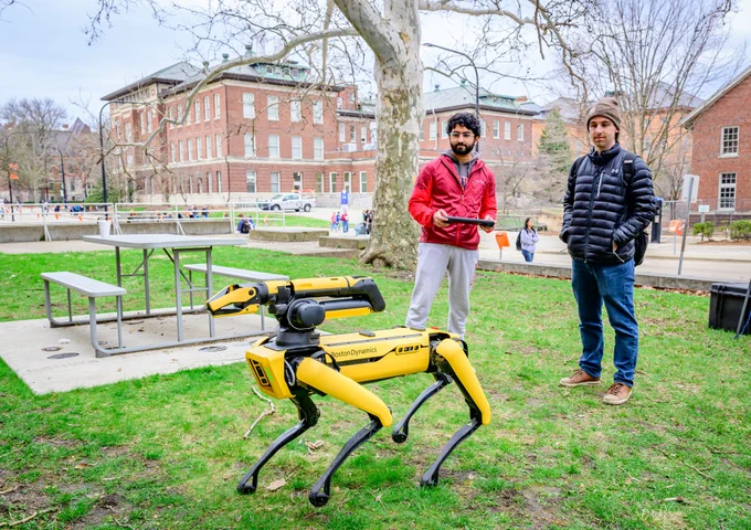 A Grainger engineering student use a remote control to make a yellow robot, which resembles an insect with four legs move on the Engineering Quad, as a spectator looks on.
