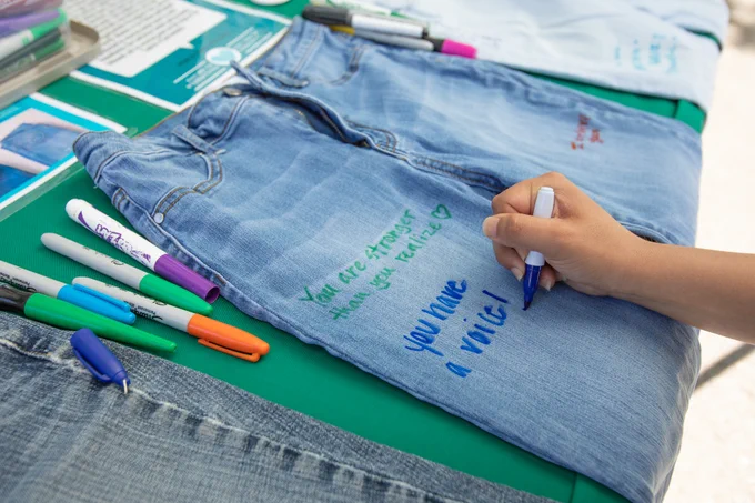 Person writing supportive messages on denim jeans for a denim day event, surrounded by colorful markers.
