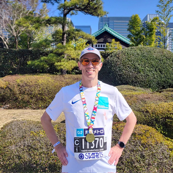 A man wearing a white running shirt with a blue logo for The Change Foundation wears a 2025 Tokyo Marathon finisher's medal around his neck. In the background are some bushes, a traditional Japanese building, and modern skyscrapers.