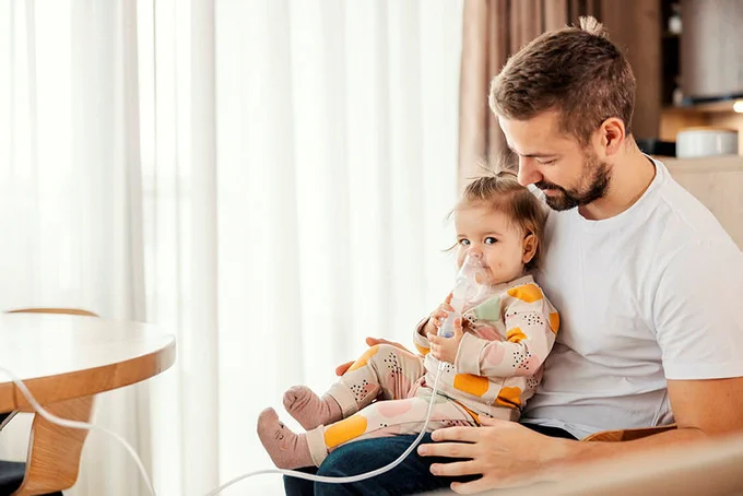 A child is inhaling medicine using a nebulizer sat on her father's knee