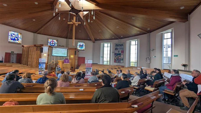 Participants of the rock art workshop listening to Helen Spencer give an introductory talk in the Faifley Parish Church, a round church building with wooden benches and wood panelled ceiling. 