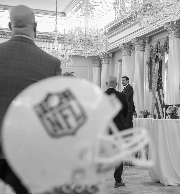 Secretary Rubio stands behind a podium adorned with the American flag in an ornate room with chandeliers and columns. In the foreground, there is an NFL helmet prominently displayed. (Official State Department photo by Freddie Everett)