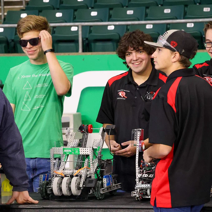 A group of competitors smile and laugh together as they prepare their robots to compete on stage.