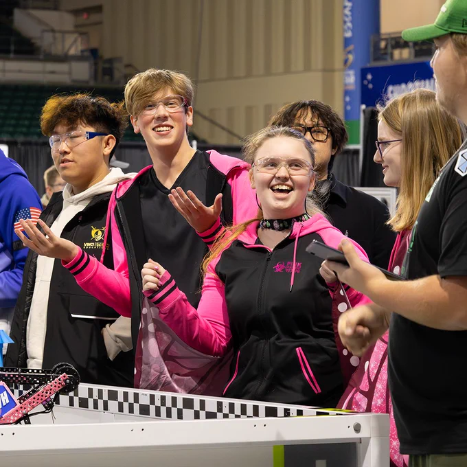 A group of students in matching outfits laugh and throw their hands into the air, directed towards competition onlookers.