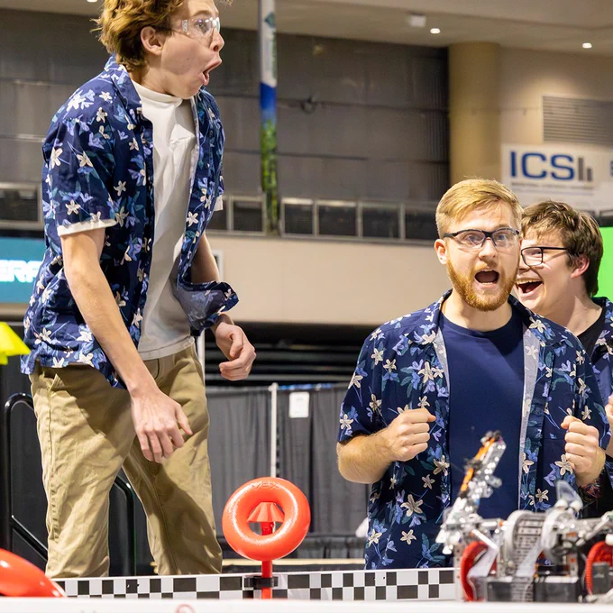 A group of robotics competitors celebrates excitedly, with one of the competitors numping several feet in the air.