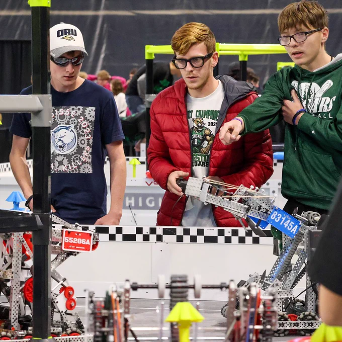 Two teams work together on the robotics playing field. They are very focused on the competition in front of them as a leading student points and directs the robot's operator for its next move.