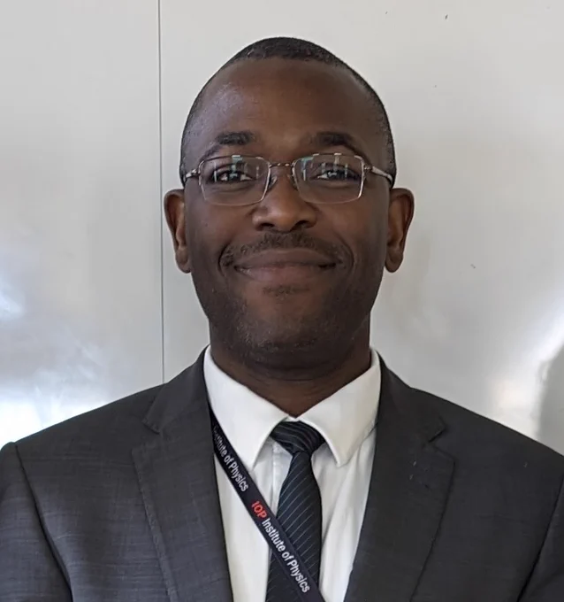 Image shows Dr Adetunmise Dada standing face-to-camera in front of a white panelled wall. He wears a white shirt with a black striped tie, grey blazer and an Institute of Physics lanyard.