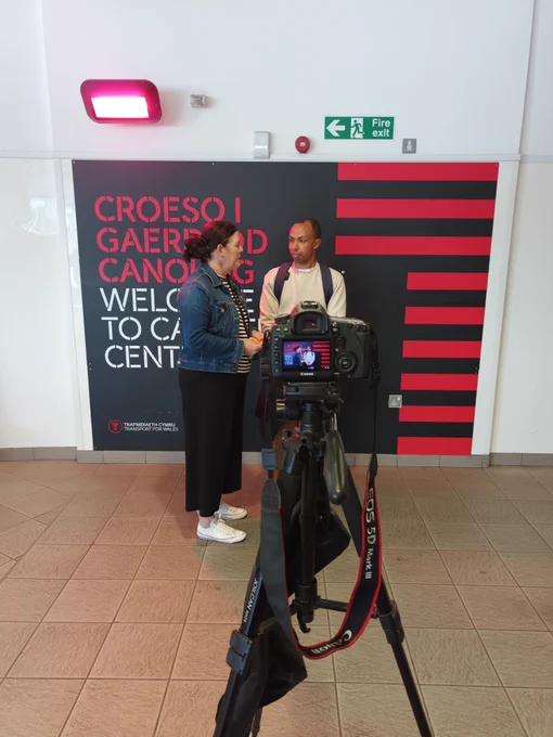 2 individuals are seen through a camera screen. They are being filmed talking in front of a sign that reads "Welcome to Cardiff Central Station".