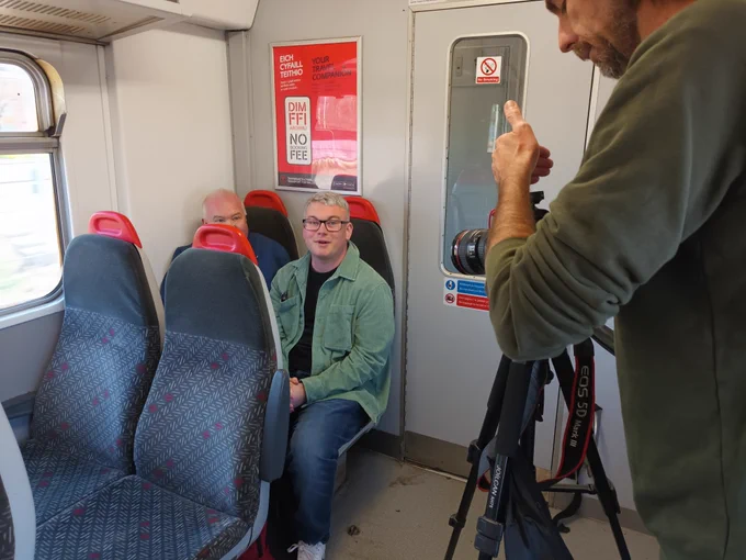 2 individuals are being filmed while they sit side by side on a train.