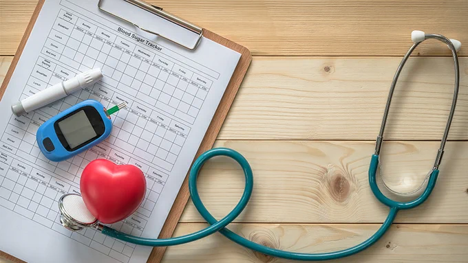 Flatlay image with a clipboard holding a blood sugar tracker document, blood testing devices, a plastic heart-shaped figure, and a stethoscope.
