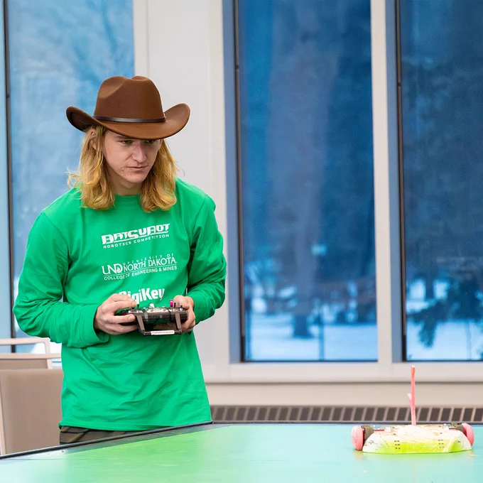 A student uses a remote control to operate a robot moving on a green battle platform. His expression is intense and he wears a brown cowboy hat.