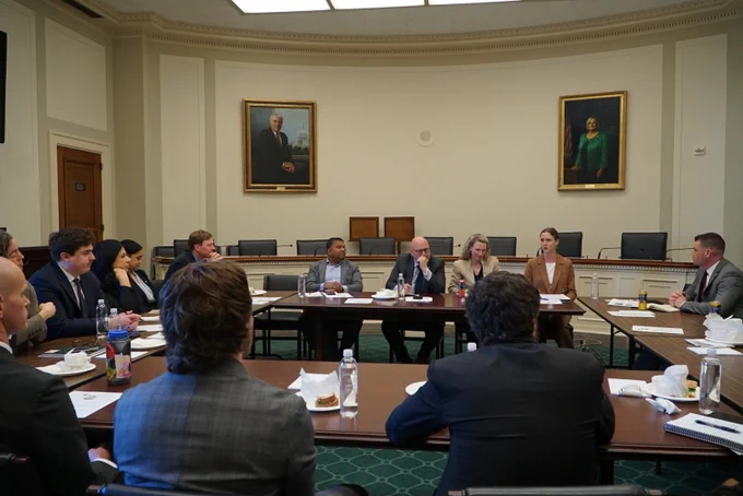 Over a dozen people sitting behind tables in a square, watching a panel of speakers, on Capitol Hill.
