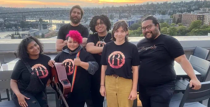 A group of 6 smiling people wearing shirts with the Rhapsody Project logo