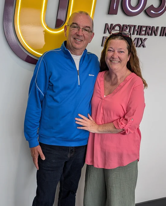 Image shows Trevor and Jennifer standing smiling together. Trevor is wearing a bright blue jacket and dark jeans and Jennifer looks lovely in a pink blouse and wide-leg trousers.