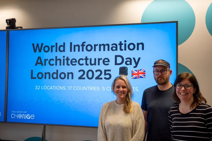 Three information architects standing in front of a large screen displaying a presentation slide which reads: “World Information Architecture Day London 2025. 32 locations, 17 countries, 5 continents.”