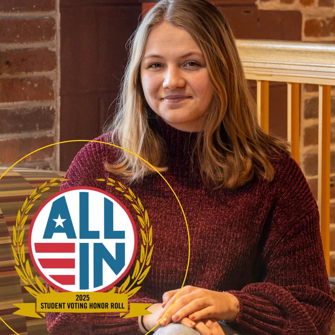 A young woman with long, wavy hair sits in a cozy setting, wearing a burgundy sweater. She is smiling and positioned near wooden seating. In the foreground, a graphic features the text "ALL IN 2025 STUDENT VOTING HONOR ROLL" enclosed within a laurel wreath.