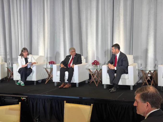 Three people seated on a stage, two men on the right and one woman on the left, dressed in business attire and engaged in a discussion.