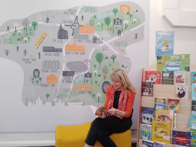 A person is sitting on a yellow couch, reading a book, in a library with a colourful wall mural of a map showing various places. Shelves with books and are visible to the right.