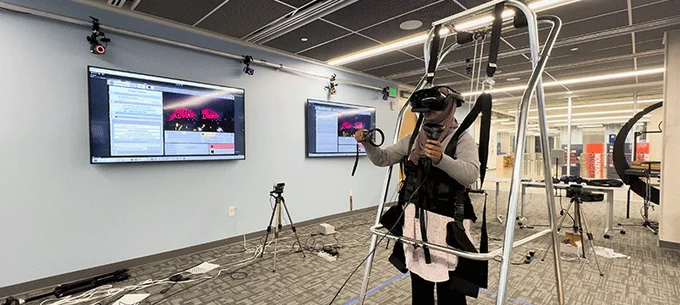 Person using a virtual reality headset and equipment in a lab setting, featuring multiple screens displaying code and graphics, and various technological devices around the room.