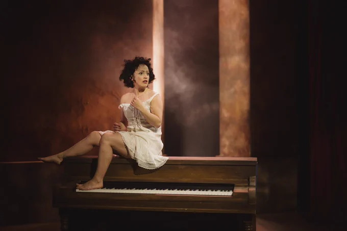 A woman - Edith Piaf - short black hair, in a full white, sits on top of a piano looking in to the distance.