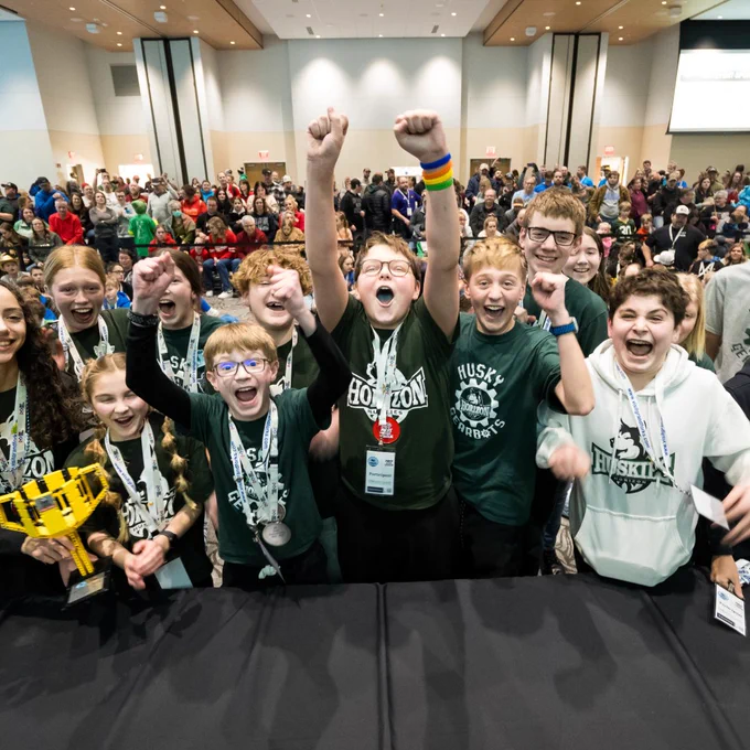A very large group of kids celebrates excitedly, many throwing their hands in the air for joy. A team in the front holds a large trophy made of LEGO bricks.