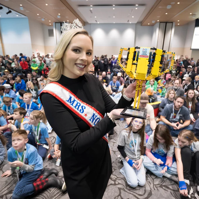 A woman wearing a 'Mrs. North Dakota' sash and a crown presents a trophy made out of LEGO bricks proudly with a smile in front of a ballroom full of kids.