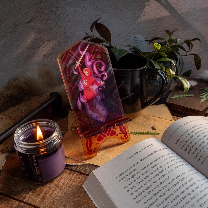 The Alanna Acrylic Phone Standee is placed on a warm table in a cottage, with an open copy of The Woman Who Rides Like a Man, the Dancing Dove Mug, and a lit Briarwick Alanna Candle.