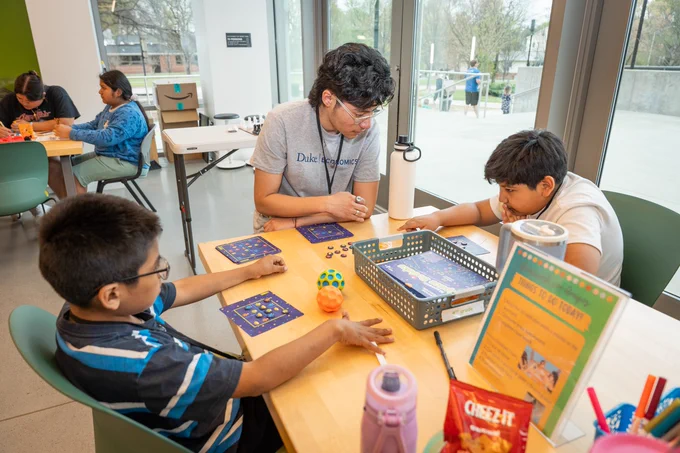 Children gather around a table in a bright community space, playing games and coloring with snacks nearby. A young adult helps two boys focused on game cards, while others join in activities across the room. Natural light from large windows fills the space with warmth and energy.