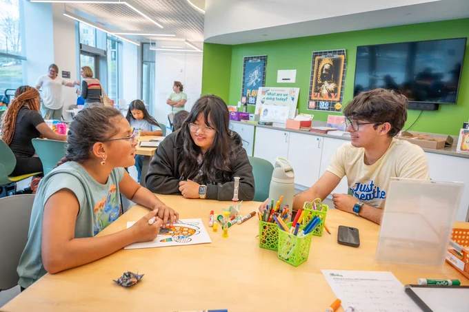 Three young people sit at a table chatting and sharing art supplies. One draws while the others talk. A bright green wall with art and a TV screen decorates the background, where more people are seen joining in creative activities around the room.